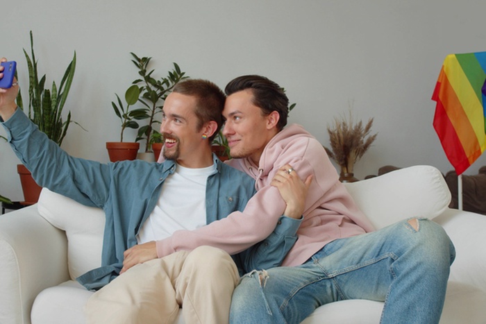 Waist up portrait of young same sex couple embracing by window at home and smiling carefree